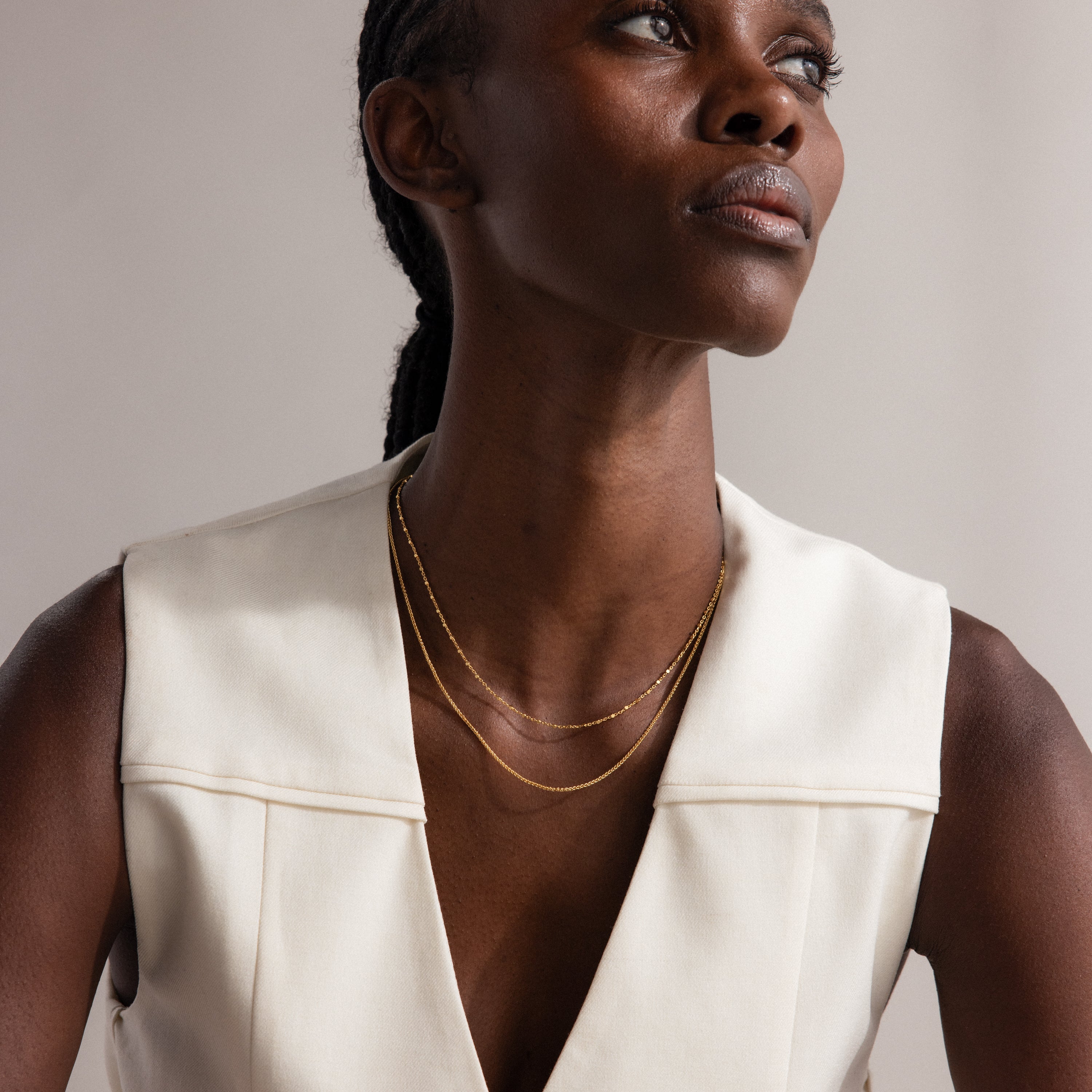 Woman in a sleeveless cream top wears the Woven Chain Necklace layered with a charm necklace, gazing upward with a neutral expression.