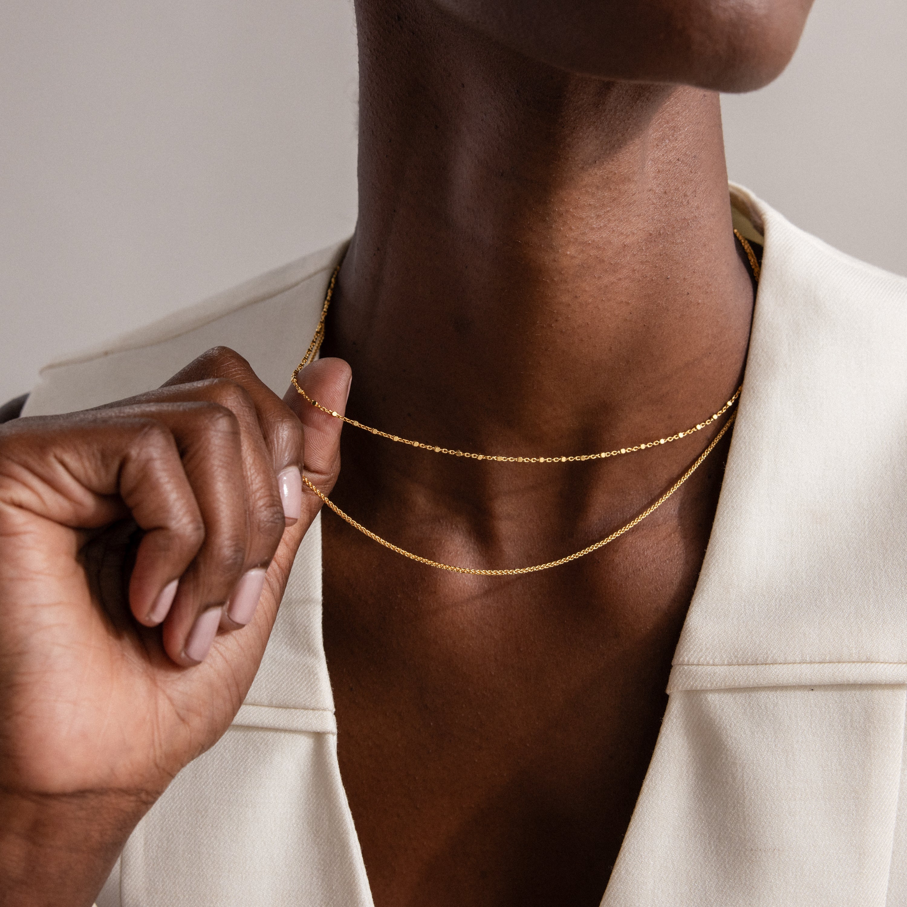 A person wearing a cream-colored collared top layers necklaces, including the Woven Chain Necklace, and holds one necklace.