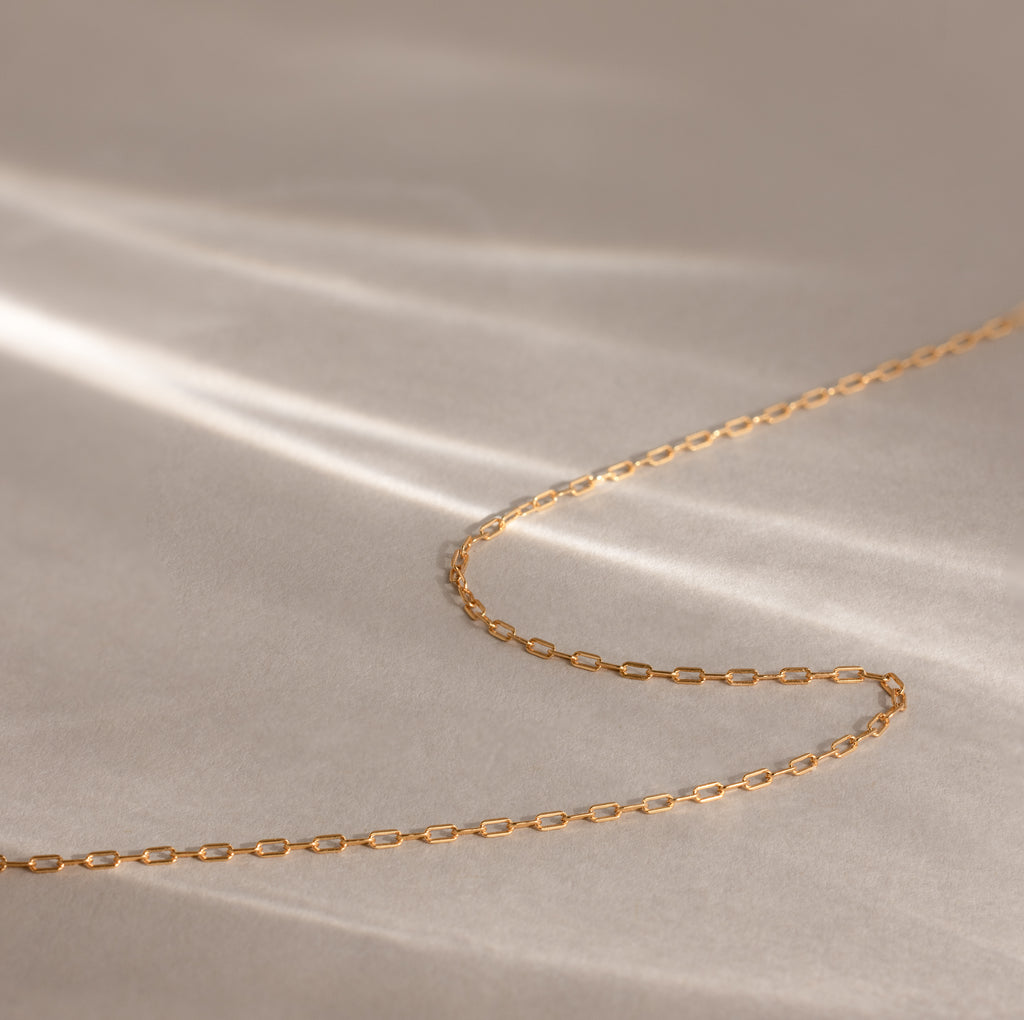 The Dainty Paperclip Necklace in 18K Gold rests on a soft beige surface with gentle light and shadow, highlighting its modern, minimalist style—perfect for layering.