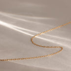The Dainty Paperclip Necklace in 18K Gold rests on a soft beige surface with gentle light and shadow, highlighting its modern, minimalist style—perfect for layering.
