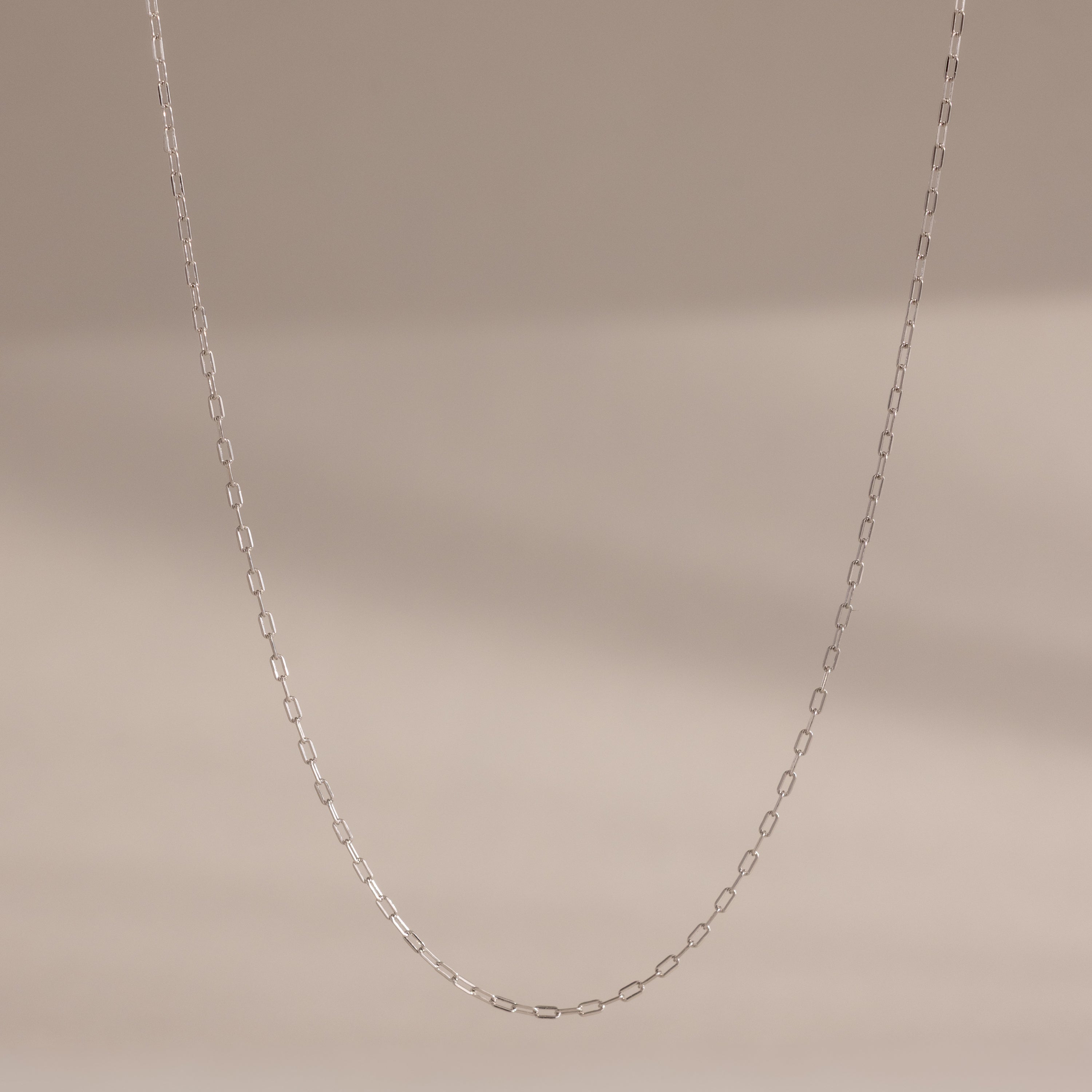 The Dainty Paperclip Necklace in Sterling Silver features small oval links and is shown against a neutral beige background—a perfect layering piece for modern jewelry lovers.