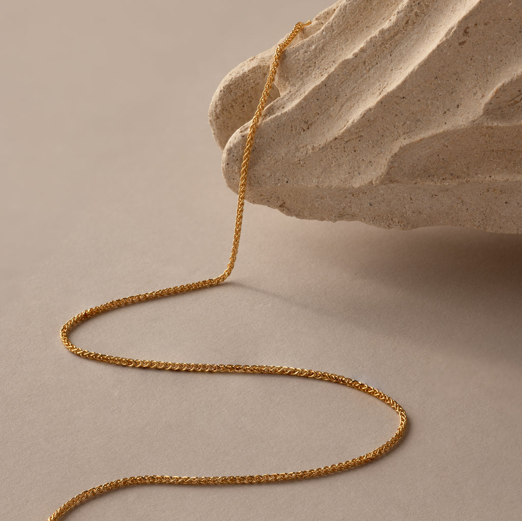 The Woven Chain Necklace in 18K Gold drapes over a textured beige stone, its interlaced links gleaming against a neutral background.