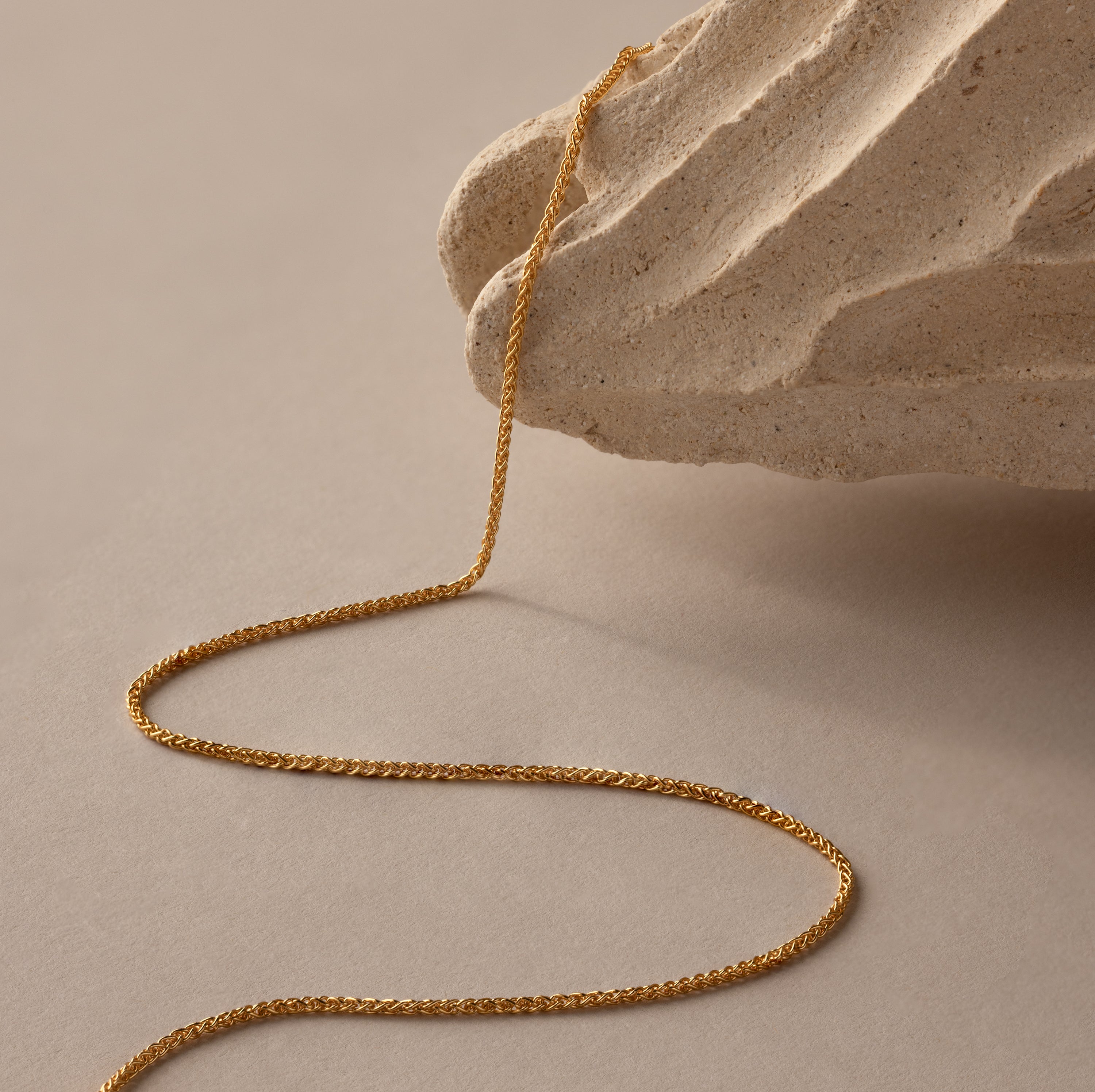 The Woven Chain Necklace in 18K Gold drapes over a textured beige stone, its interlaced links gleaming against a neutral background.