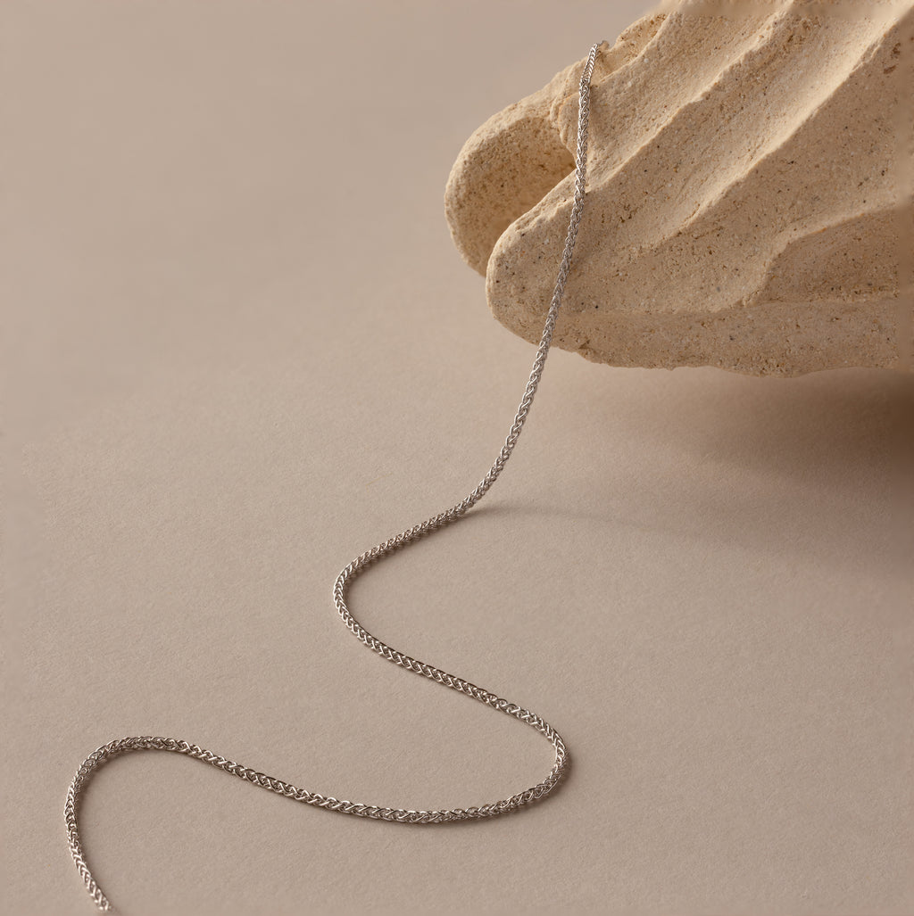 The Woven Chain Necklace in Sterling Silver is draped over a textured beige stone on a neutral background, ideal for layering with your favorite jewelry.