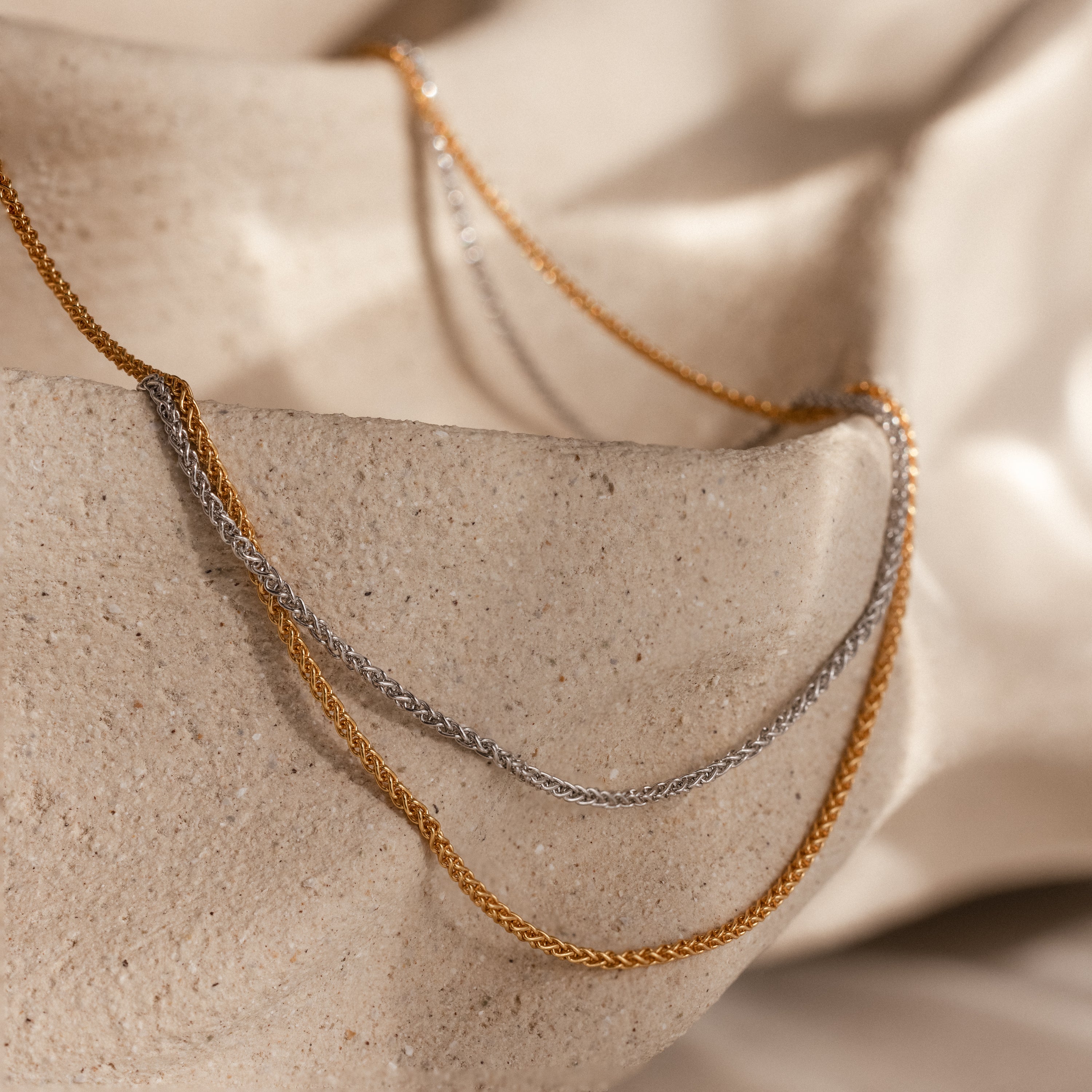 The Woven Chain Necklace draped over textured beige stone, bathed in soft, natural lighting.