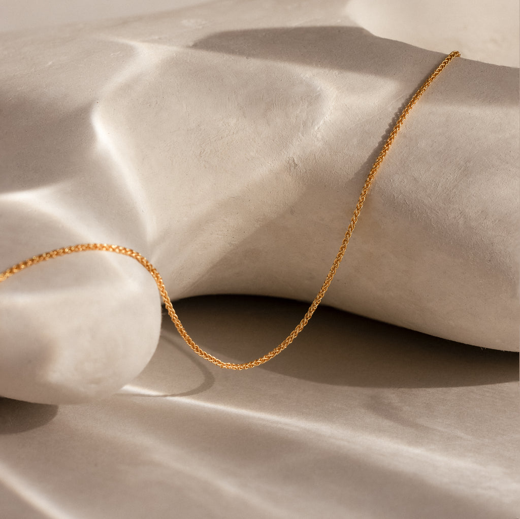 The Woven Chain Necklace in 18K Gold, featuring interlaced links, is displayed draped over a smooth, light-colored sculpted surface in soft lighting.