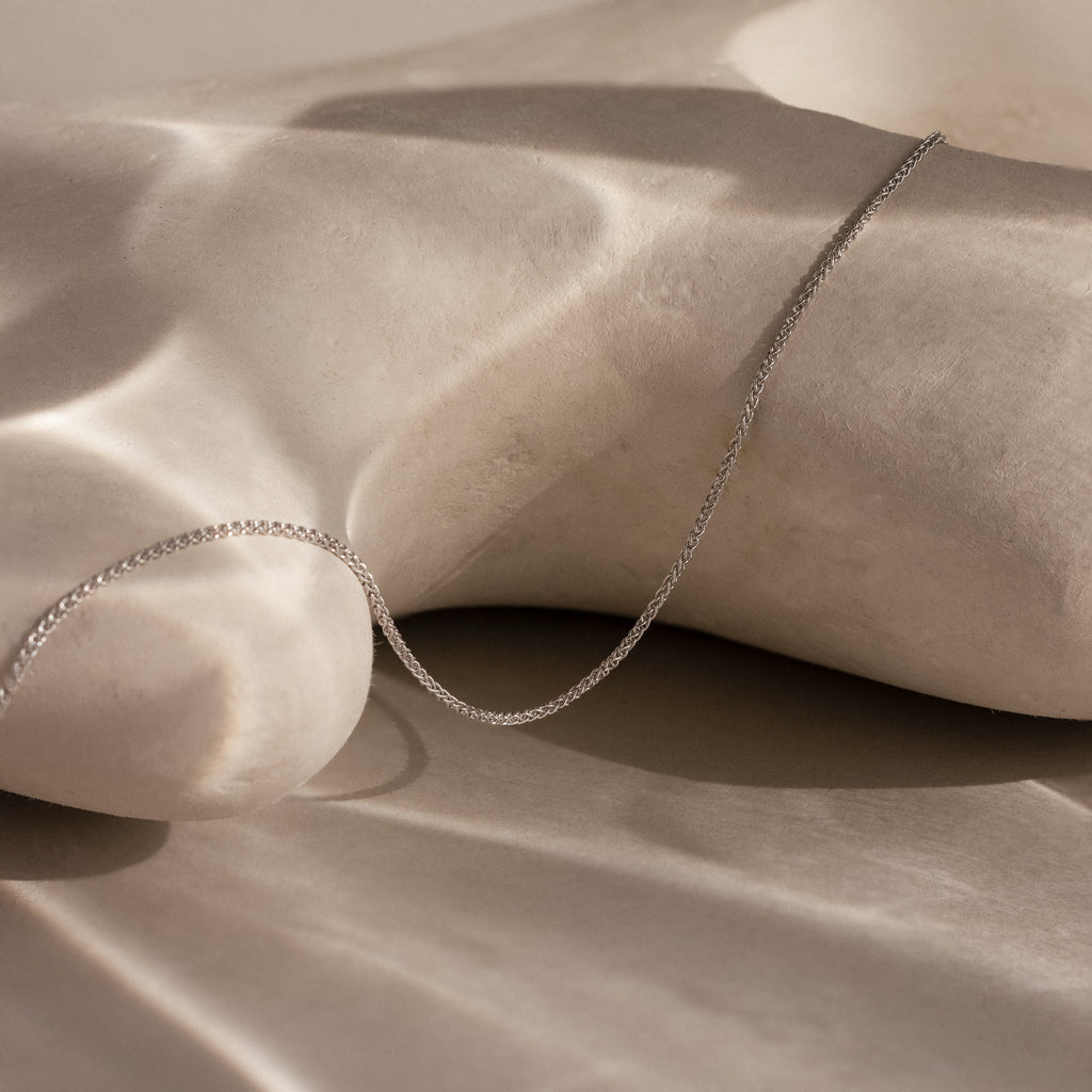 The Woven Chain Necklace in Sterling Silver is displayed on a smooth, light-hued sculpted surface with soft shadows.