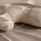 The Woven Chain Necklace in Sterling Silver is displayed on a smooth, light-hued sculpted surface with soft shadows.