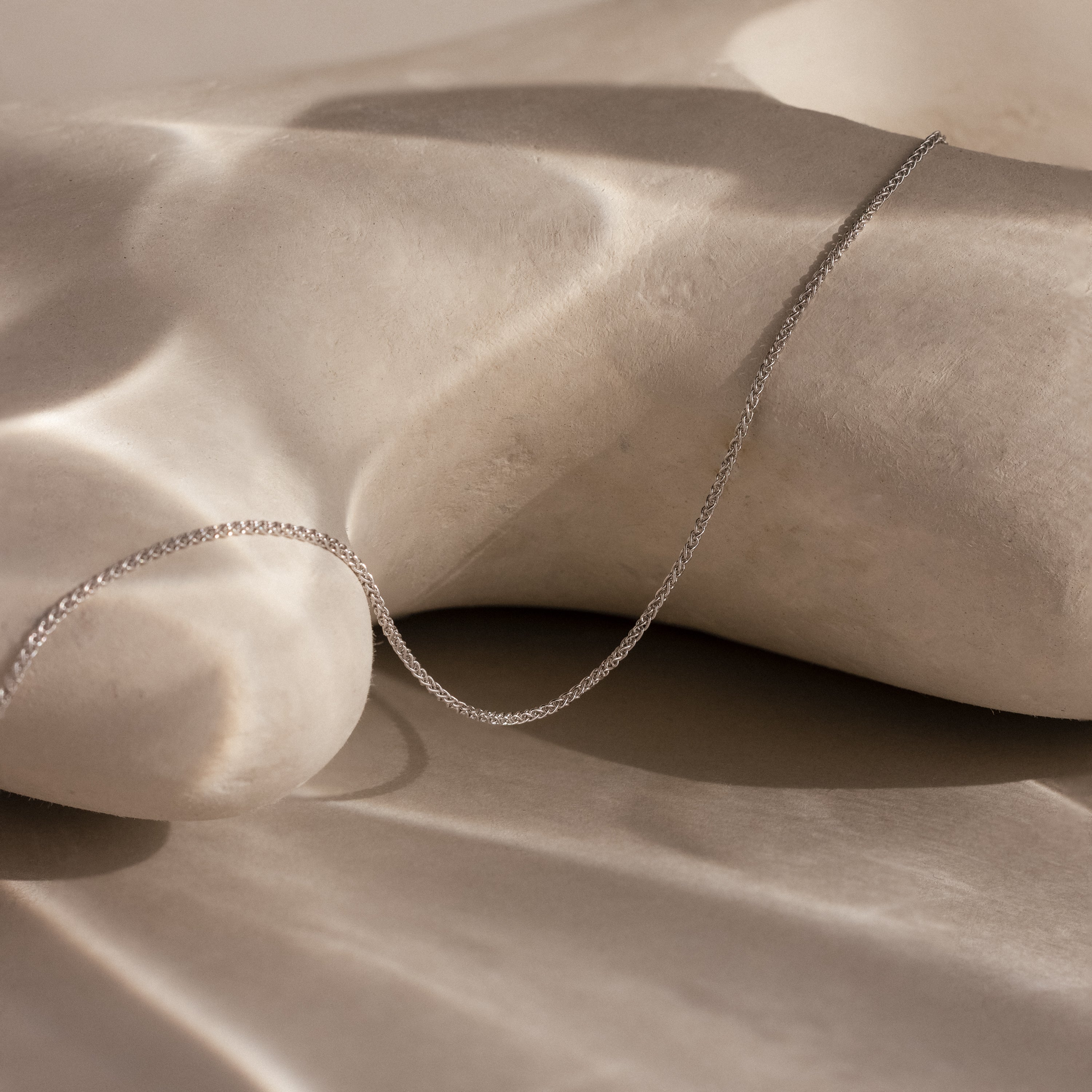 The Woven Chain Necklace in Sterling Silver is displayed on a smooth, light-hued sculpted surface with soft shadows.