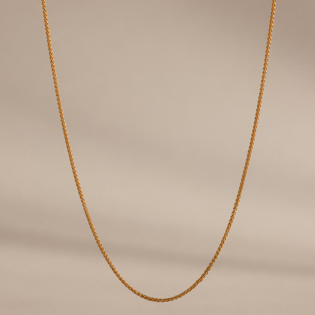 The Woven Chain Necklace in 18K Gold, featuring interlaced links, is displayed against a beige background.