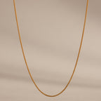 The Woven Chain Necklace in 18K Gold, featuring interlaced links, is displayed against a beige background.
