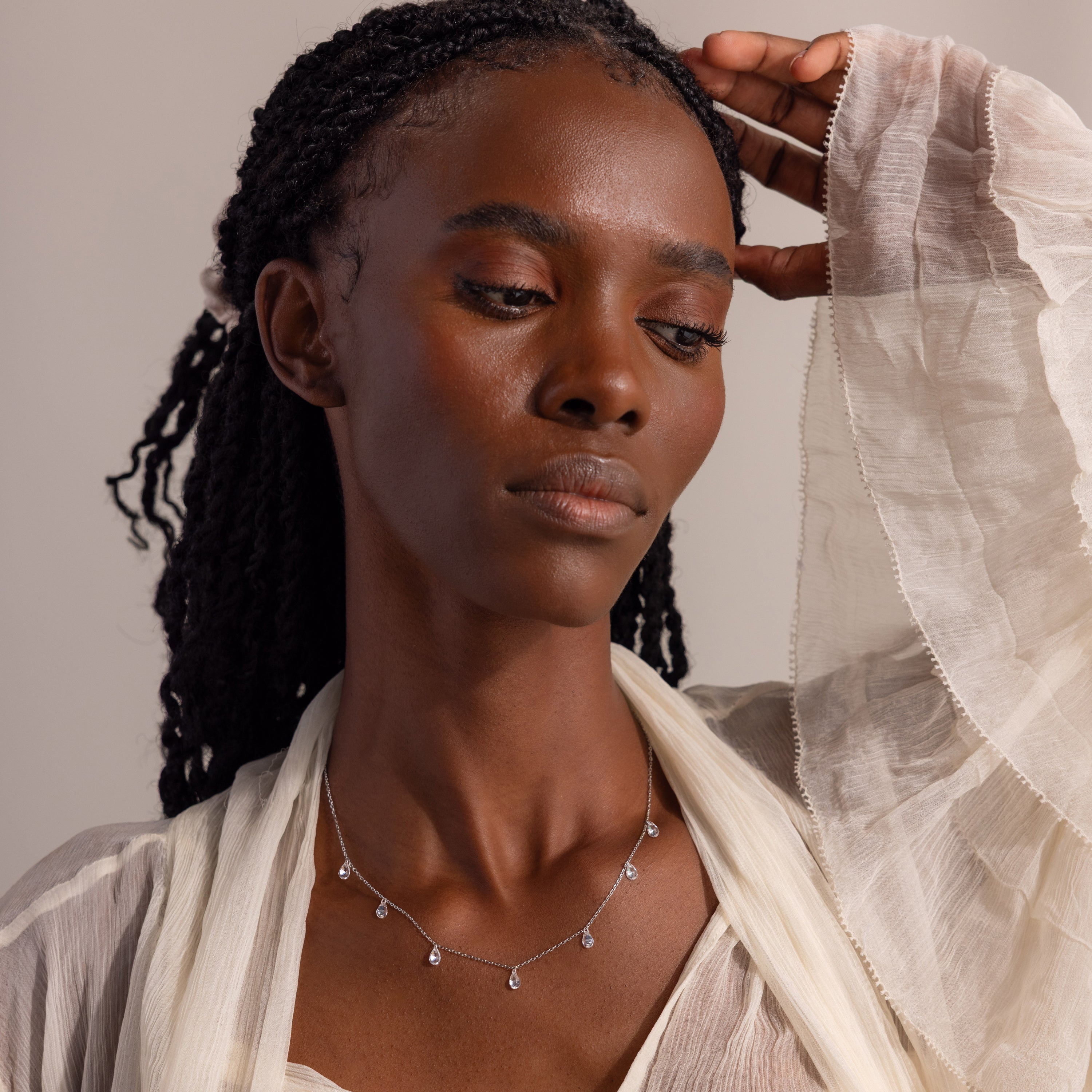 Model wearing a sheer, flowing cream blouse gazes thoughtfully downward while showcasing a delicate silver necklace adorned with evenly spaced teardrop-shaped diamond charms.