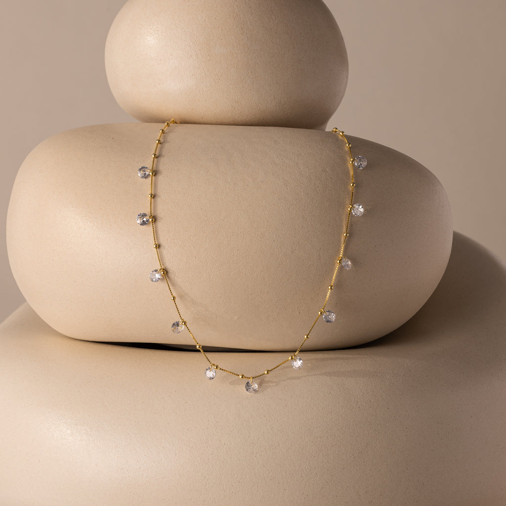 The Satellite Diamond Necklace in 18K Gold, featuring delicate crystal charms, is displayed on smooth, stacked beige stones with a neutral backdrop.