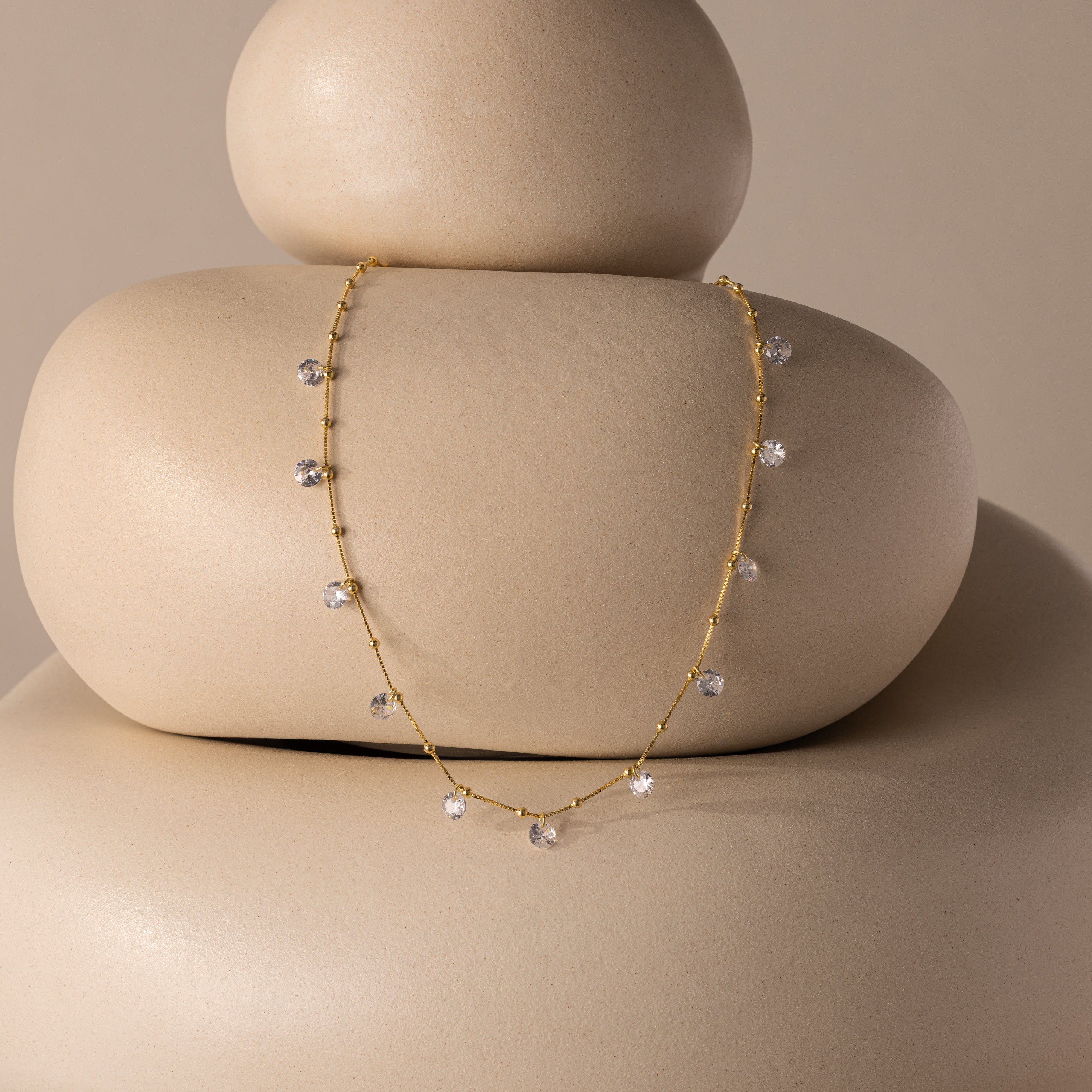 The Satellite Diamond Necklace in 18K Gold, featuring delicate crystal charms, is displayed on smooth, stacked beige stones with a neutral backdrop.