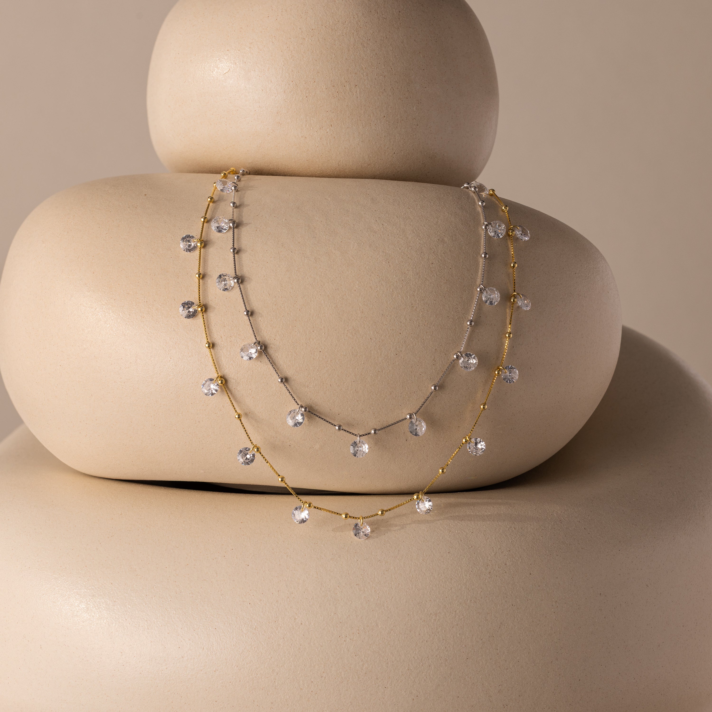 Two dainty necklaces—one silver, one gold—rest on smooth stones, each reflecting the elegant brilliance of the Satellite Diamond Necklace.