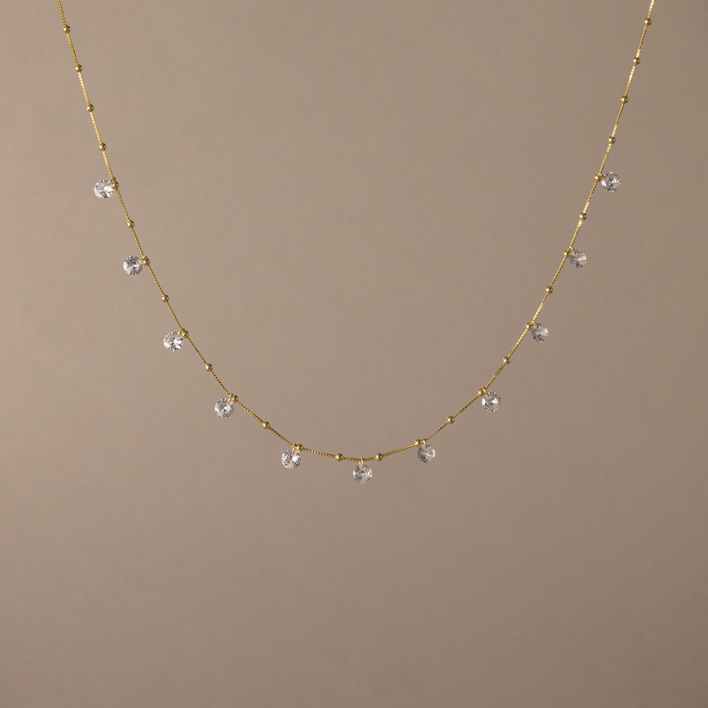 The Satellite Diamond Necklace in 18K Gold features small, evenly spaced clear crystal beads, displayed on a plain beige background.