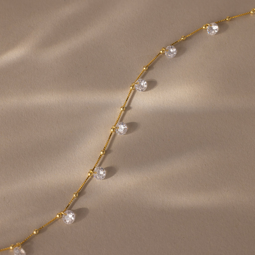 Satellite Diamond Necklace in 18K Gold, featuring spaced round clear gemstones on a beige textured background and softly lit by sunlight. This dainty piece adds delicate sparkle to any look.