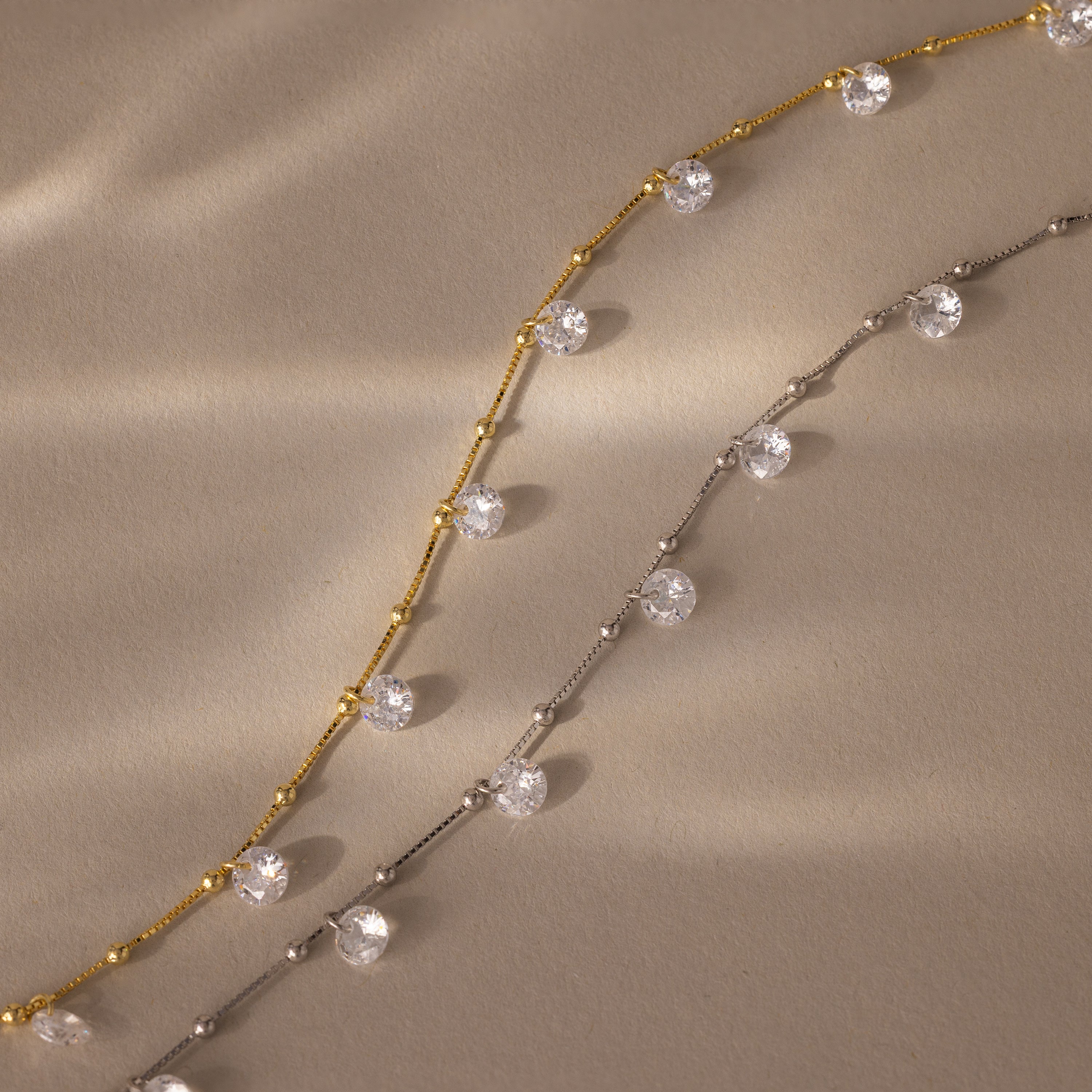 A gold and a silver chain, each featuring clear round gemstones, are displayed on a beige surface—ideal as dainty necklaces or for layering with your favorite Satellite Diamond Necklace.