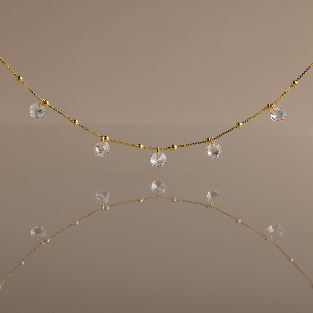 The Satellite Diamond Necklace in 18K Gold features delicate, small clear crystal drops inspired by a classic diamond design, beautifully reflected on a smooth, neutral-colored surface.