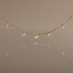 The Satellite Diamond Necklace in 18K Gold features delicate, small clear crystal drops inspired by a classic diamond design, beautifully reflected on a smooth, neutral-colored surface.