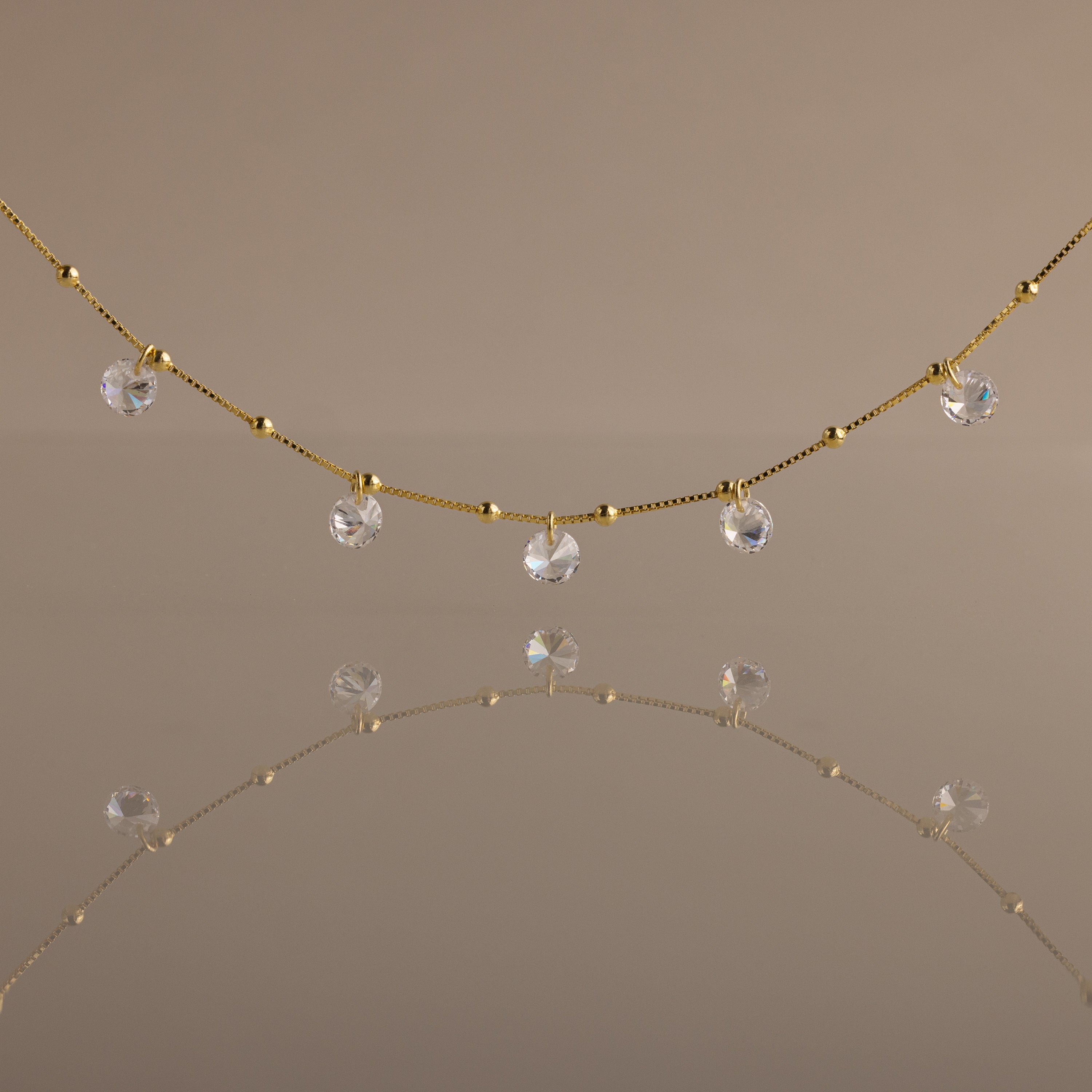 The Satellite Diamond Necklace in 18K Gold features delicate, small clear crystal drops inspired by a classic diamond design, beautifully reflected on a smooth, neutral-colored surface.