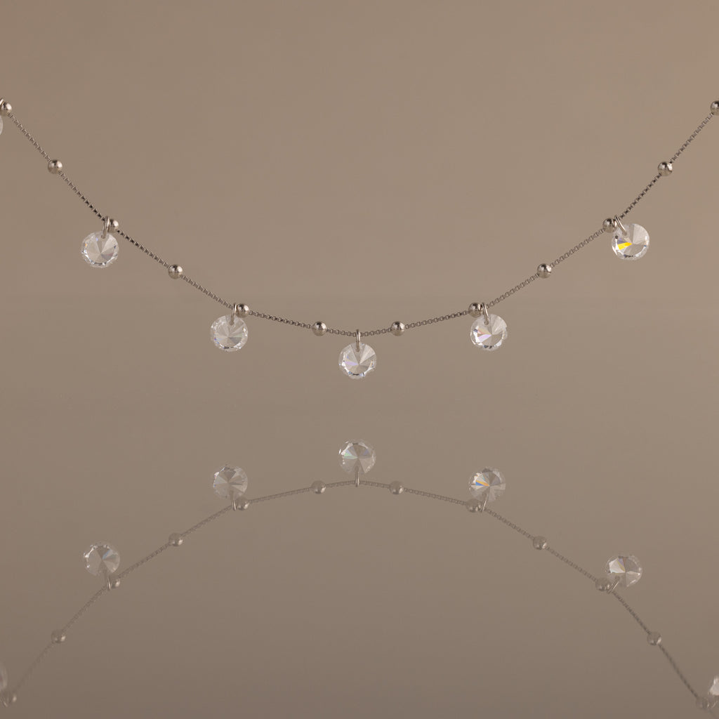 Satellite Diamond Necklace featuring small clear crystal beads, shown reflected on a smooth, neutral surface.