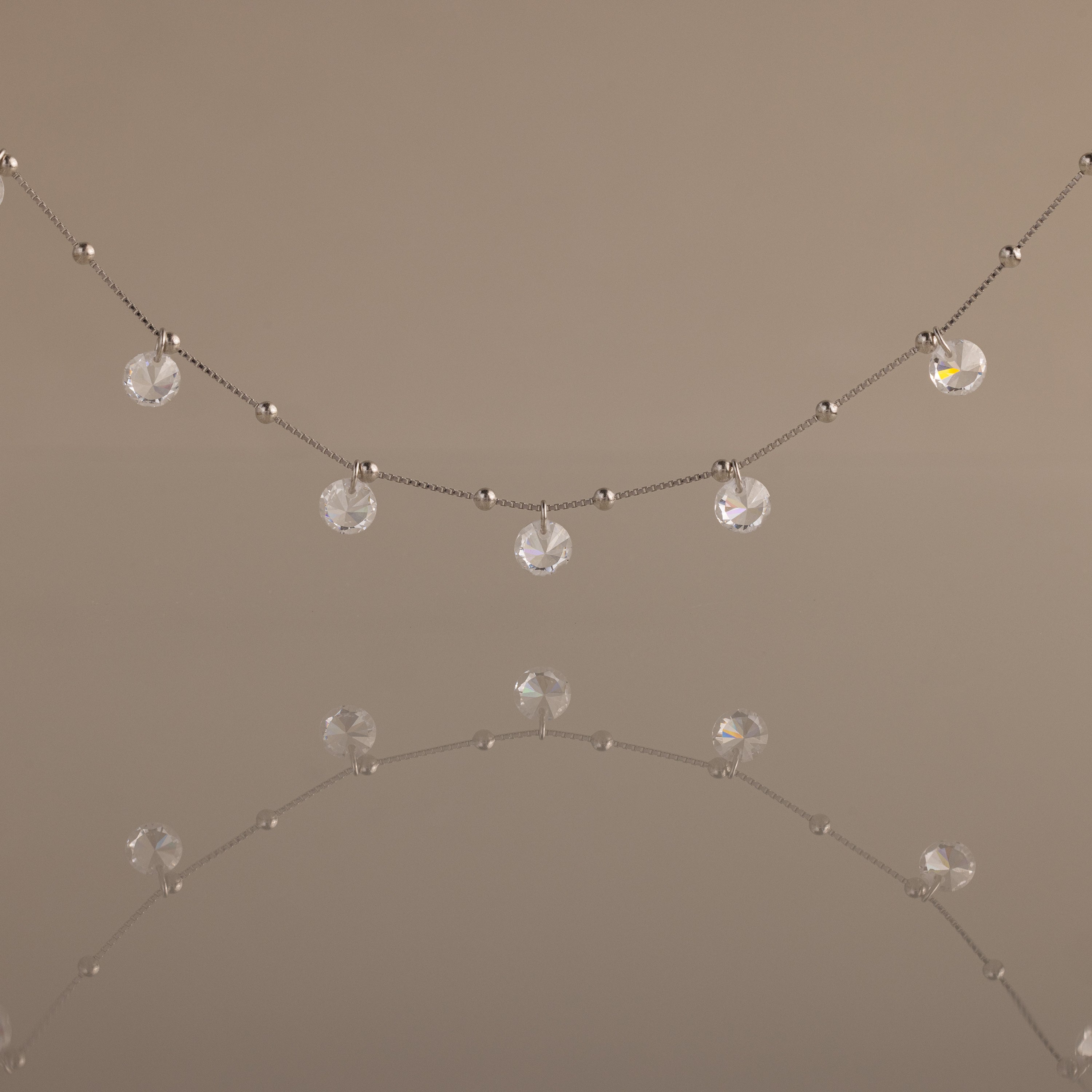 Satellite Diamond Necklace featuring small clear crystal beads, shown reflected on a smooth, neutral surface.