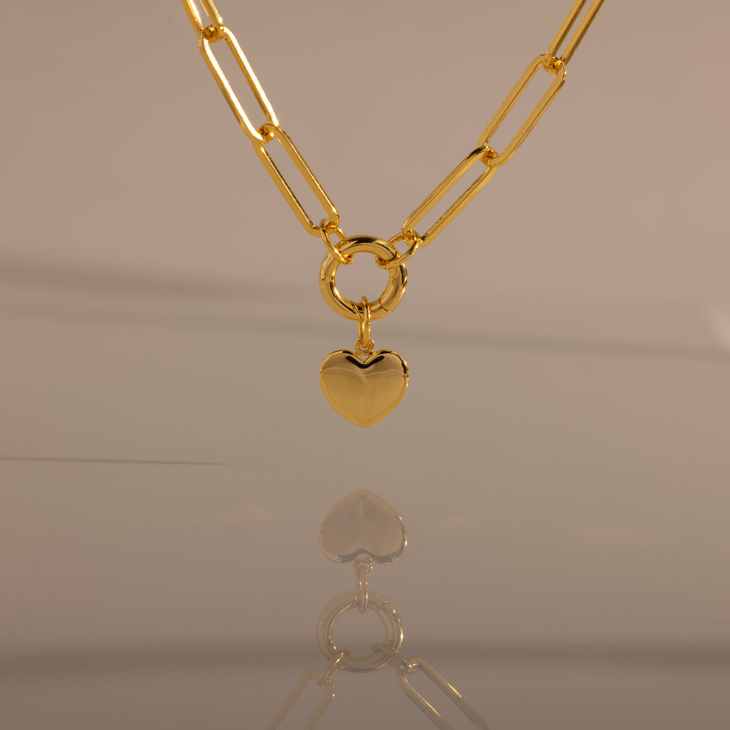 The Paperclip Heart Charm Necklace in 18K Gold features a gold paperclip chain and a heart pendant, elegantly reflected on a smooth surface against a neutral background.