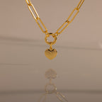 The Paperclip Heart Charm Necklace in 18K Gold features a gold paperclip chain and a heart pendant, elegantly reflected on a smooth surface against a neutral background.