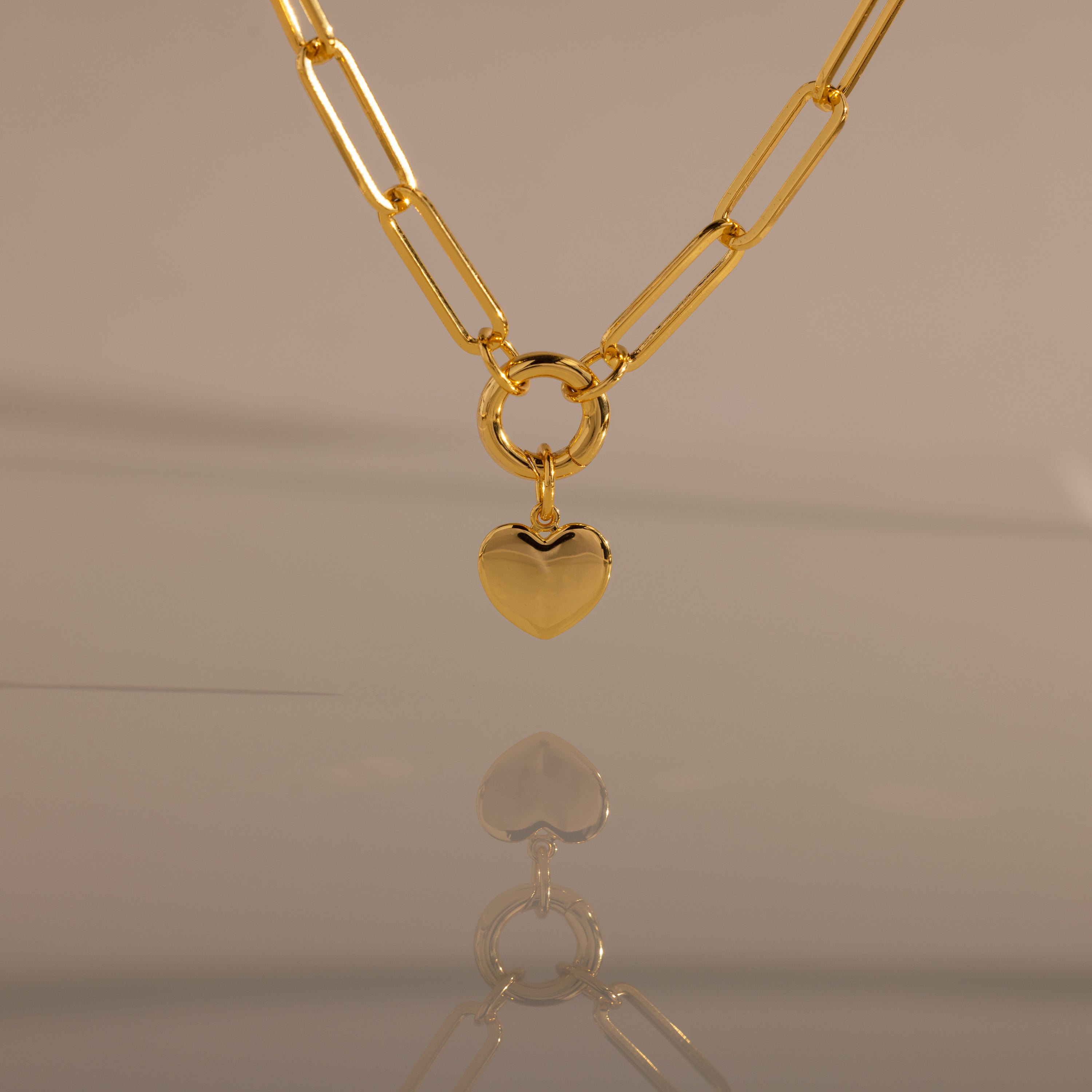 The Paperclip Heart Charm Necklace in 18K Gold features a gold paperclip chain and a heart pendant, elegantly reflected on a smooth surface against a neutral background.