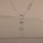 The Paperclip Heart Charm Necklace in Silver is displayed on a smooth surface, its heart pendant reflected against a neutral background.