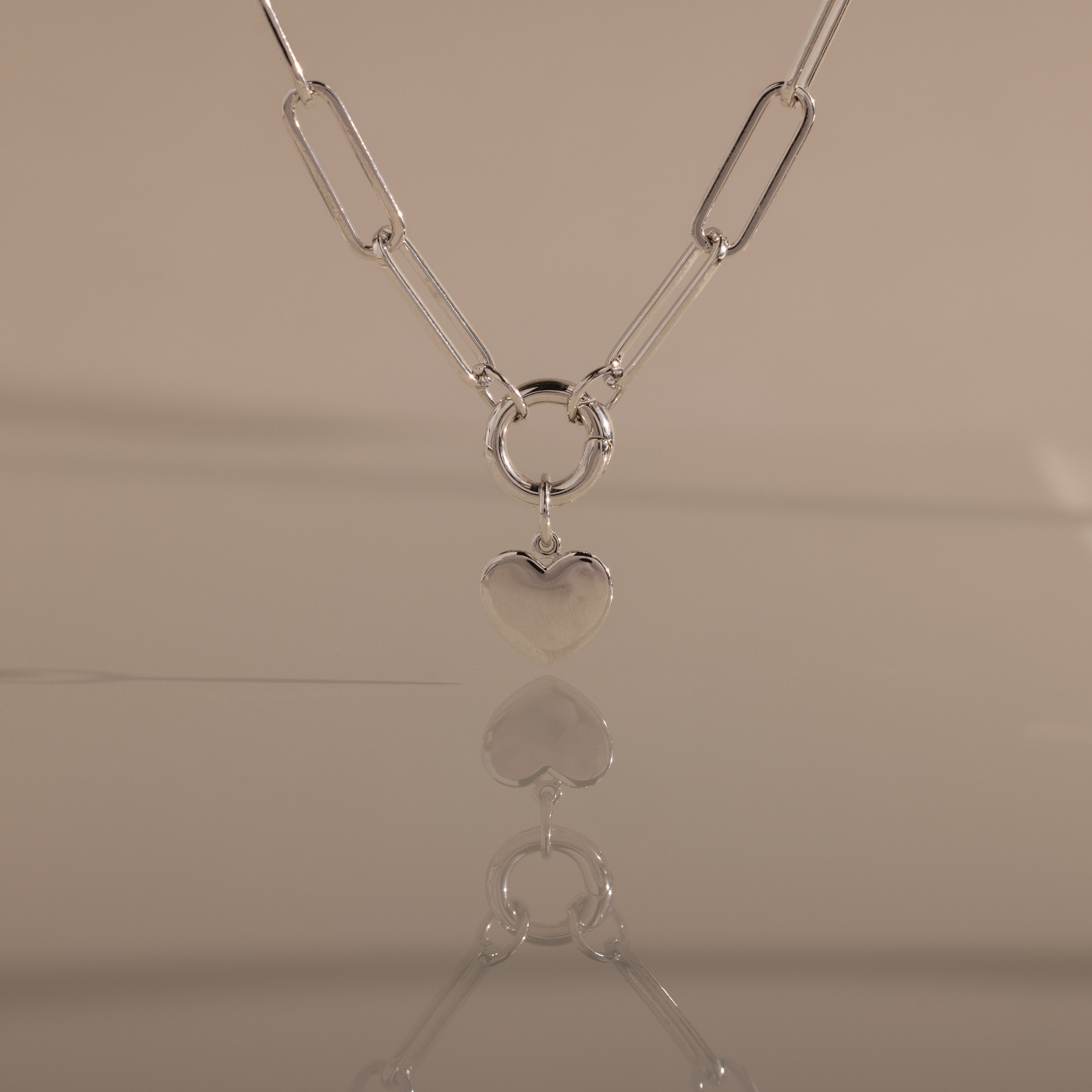 The Paperclip Heart Charm Necklace in Silver is displayed on a smooth surface, its heart pendant reflected against a neutral background.