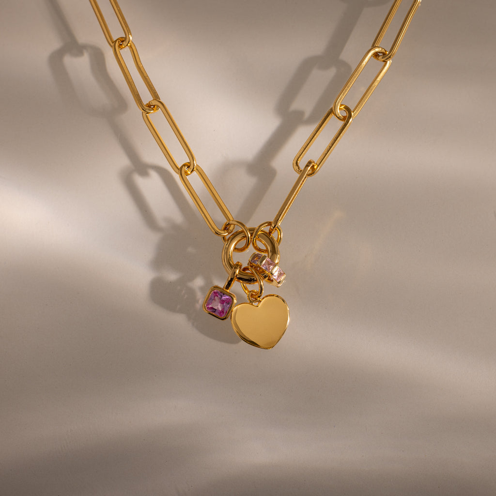 The Paperclip Heart Charm Necklace in 18K Gold features a heart pendant and two small gemstone charms, styled elegantly on a neutral background.