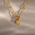 The Paperclip Heart Charm Necklace in 18K Gold features a heart pendant and two small gemstone charms, styled elegantly on a neutral background.