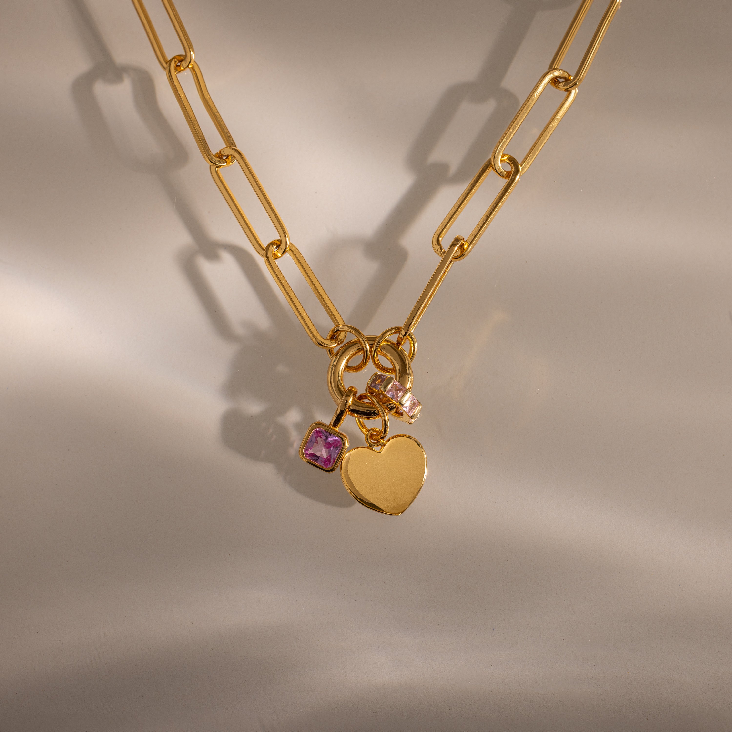 The Paperclip Heart Charm Necklace in 18K Gold features a heart pendant and two small gemstone charms, styled elegantly on a neutral background.