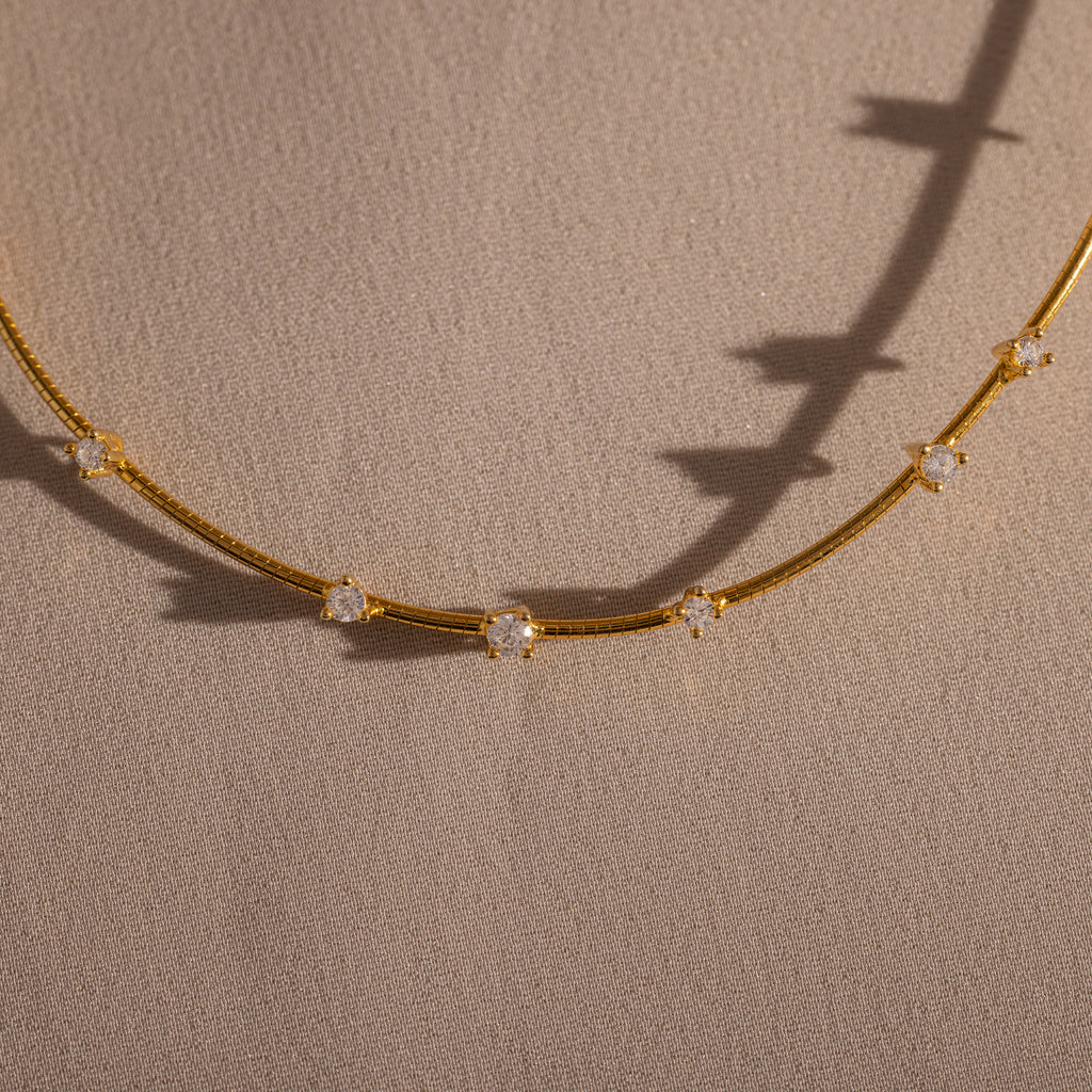 The Sprinkle Diamond Necklace in 18K Gold features a delicate chain adorned with evenly spaced sparkling stones, shown resting on a textured beige surface.