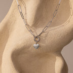 The Paperclip Heart Charm Necklace features a dainty heart pendant, elegantly displayed on a textured, abstract beige sculpture.