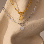 The Paperclip Heart Charm Necklace, featuring gold and silver chain styles with heart pendants, is displayed on a textured beige sculpture.