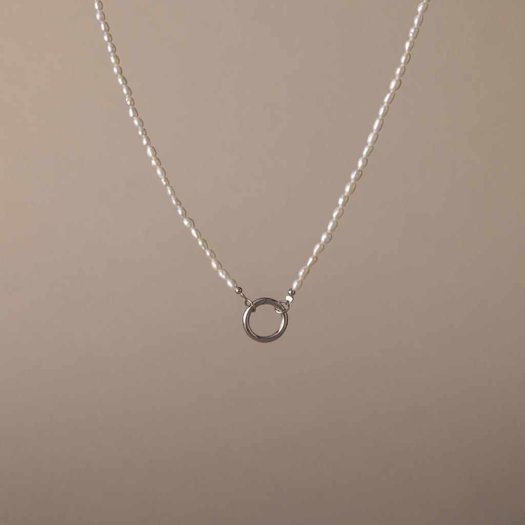 The Pearl Chain Clasp Necklace in Silver features a circular silver pendant with luminous pearls and is displayed on a plain beige background—an elegant addition to any jewelry collection.