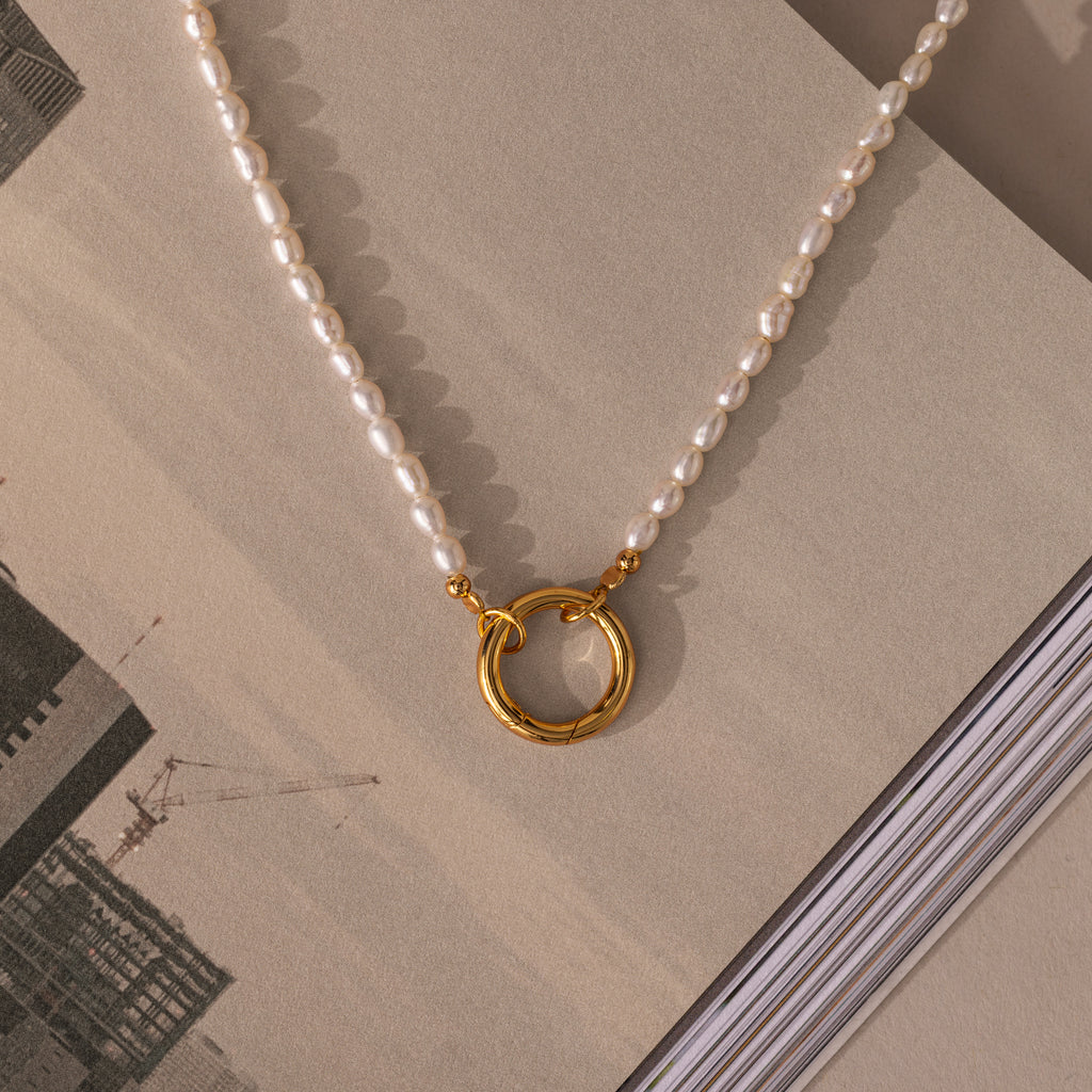 The Pearl Chain Clasp Necklace in 18K Gold, featuring a gold circular pendant, is displayed on an open book with a cityscape—an elegant and timeless addition to your jewelry collection.