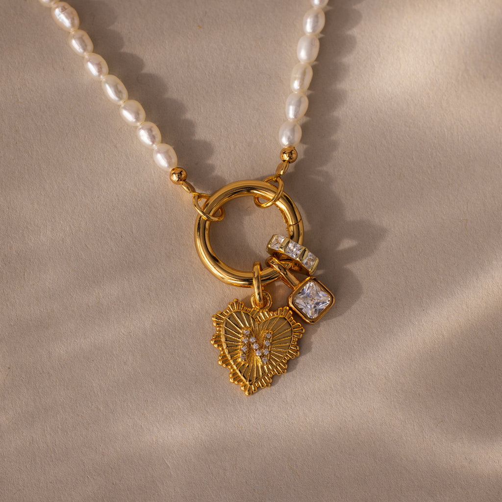 The Pearl Chain Clasp Necklace in 18K Gold, featuring gold heart and crystal charms, is a stunning addition to your jewelry collection, shown here on a beige textured surface.