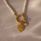 The Pearl Chain Clasp Necklace in 18K Gold, featuring gold heart and crystal charms, is a stunning addition to your jewelry collection, shown here on a beige textured surface.