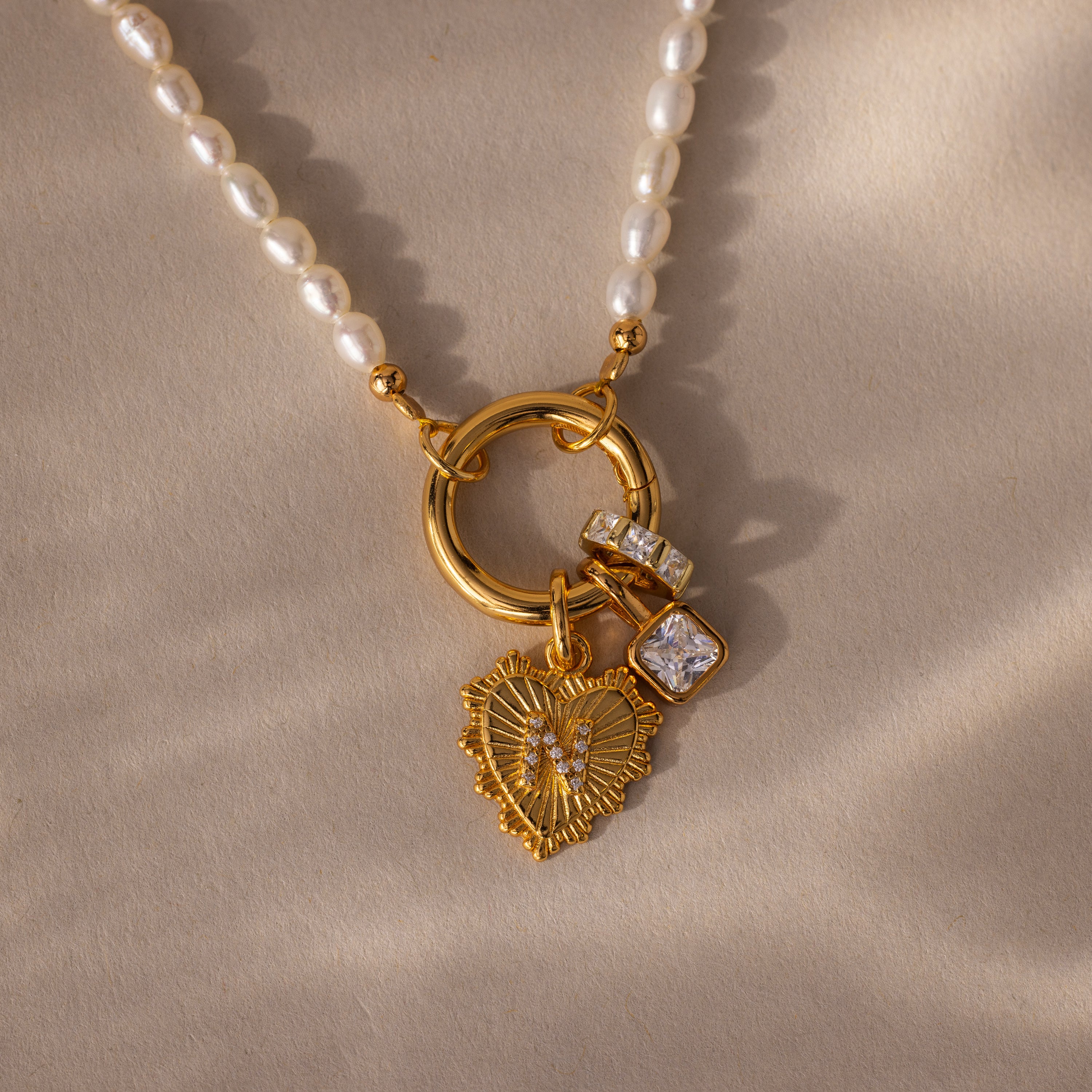 The Pearl Chain Clasp Necklace features a gold heart pendant with pearls and gemstone charms, showcased on a beige surface—a standout addition to any jewelry collection.