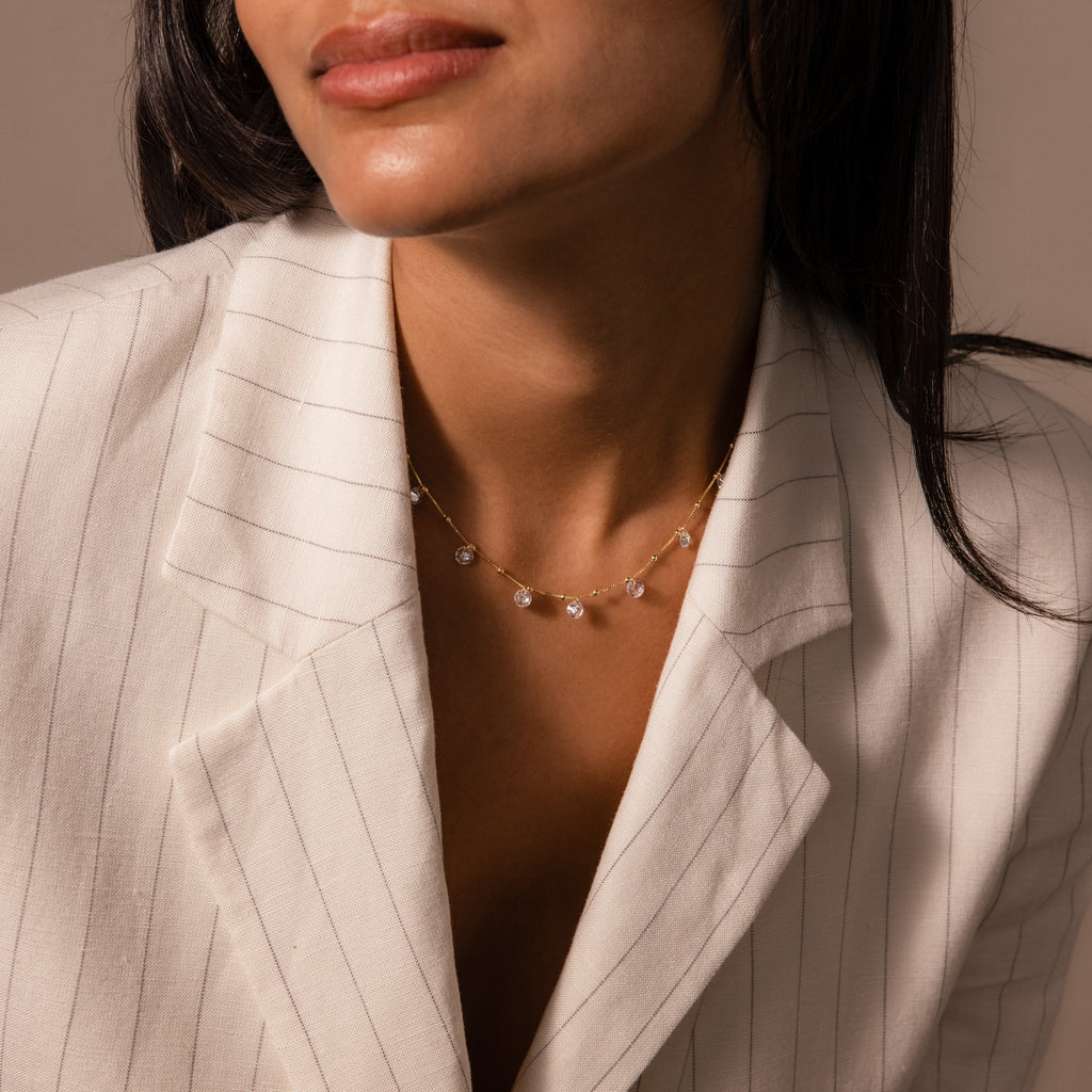 A woman in a white pinstripe blazer wears the Satellite Diamond Necklace in 18K Gold, featuring a delicate gold chain with small diamond charms.