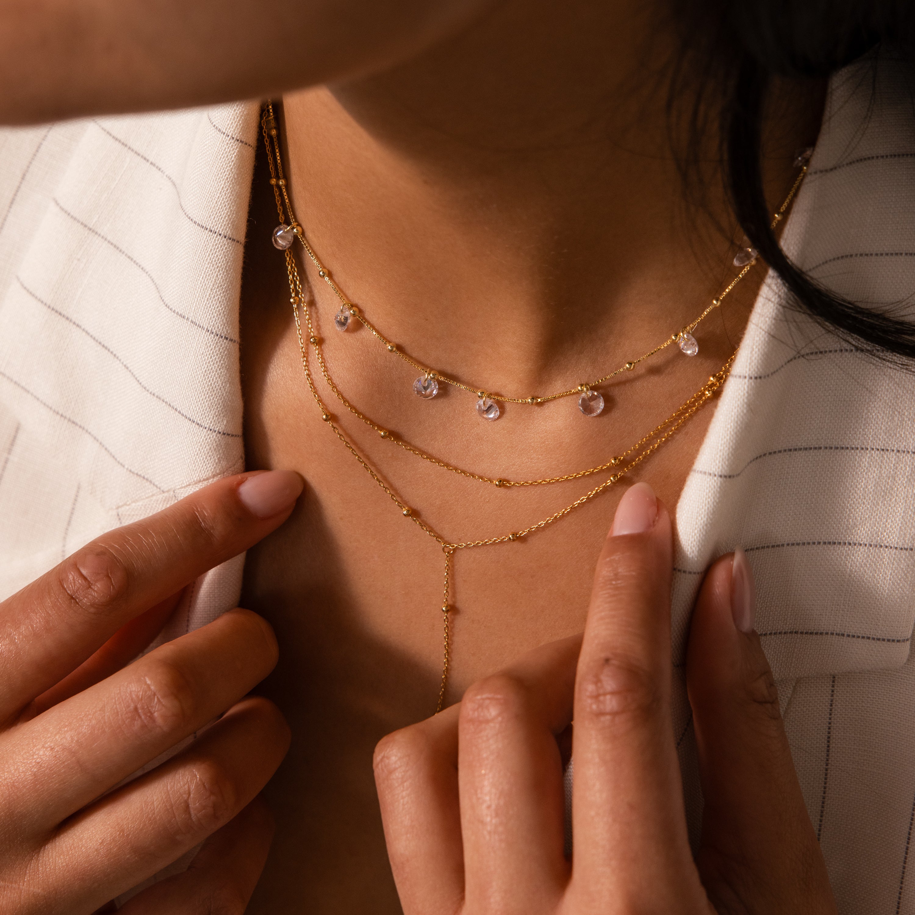 Close-up of a woman adjusting her Satellite Diamond Necklace, layered with gold pieces, while wearing a light pinstripe blazer.