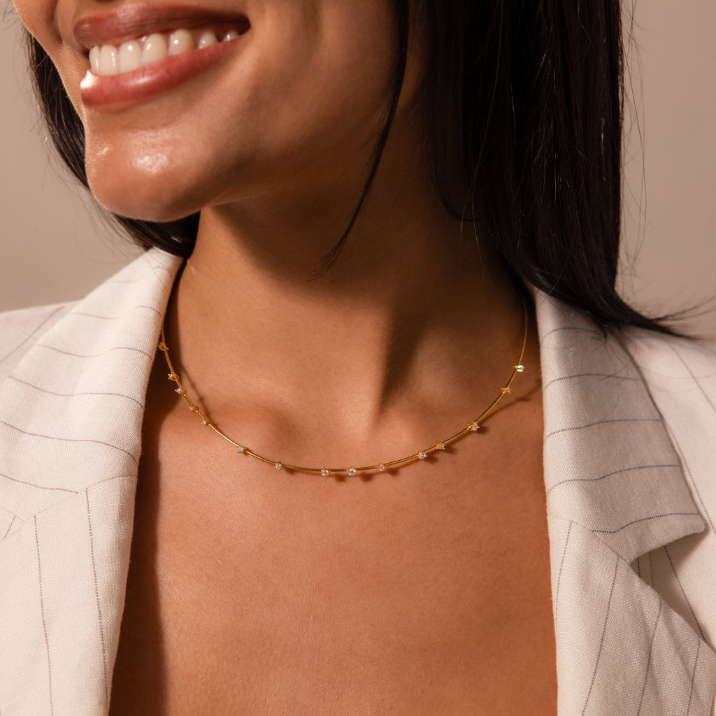 A woman in a cream blazer smiles, wearing a delicate chain layering necklace and the Sprinkle Diamond Necklace in 18K Gold adorned with small charms.