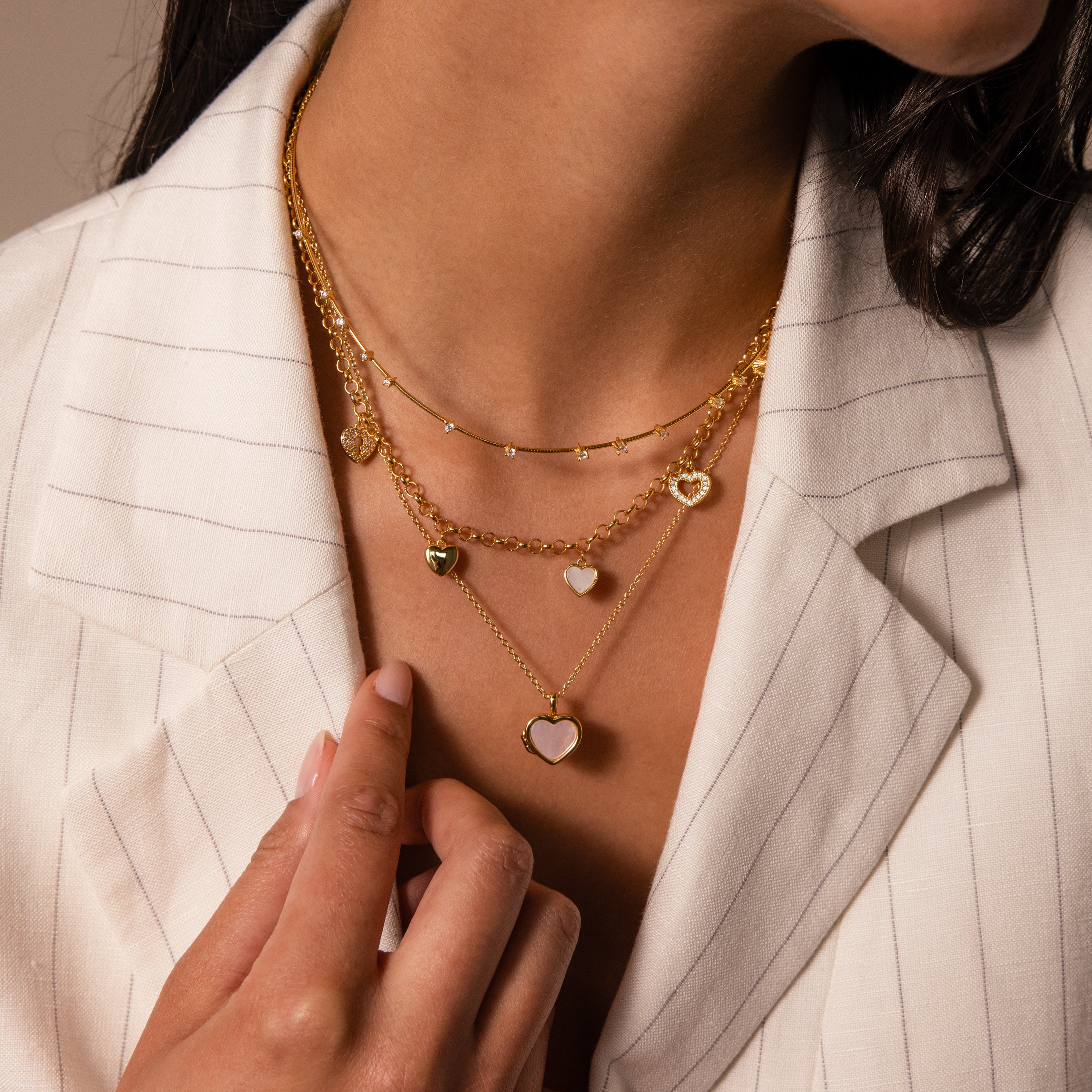 Woman in a cream pinstripe blazer wearing layered gold chains with heart-shaped pendants, including the Sprinkle Diamond Necklace in 18K Gold.