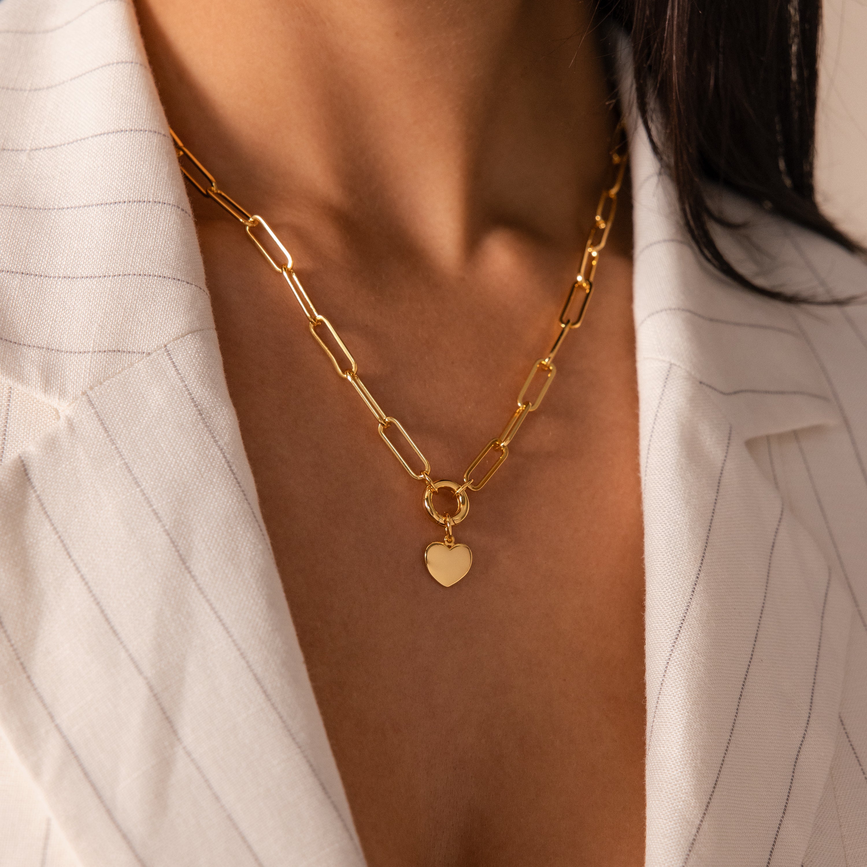 A woman wears the Paperclip Heart Charm Necklace, featuring a heart pendant, over a white pinstripe blazer.