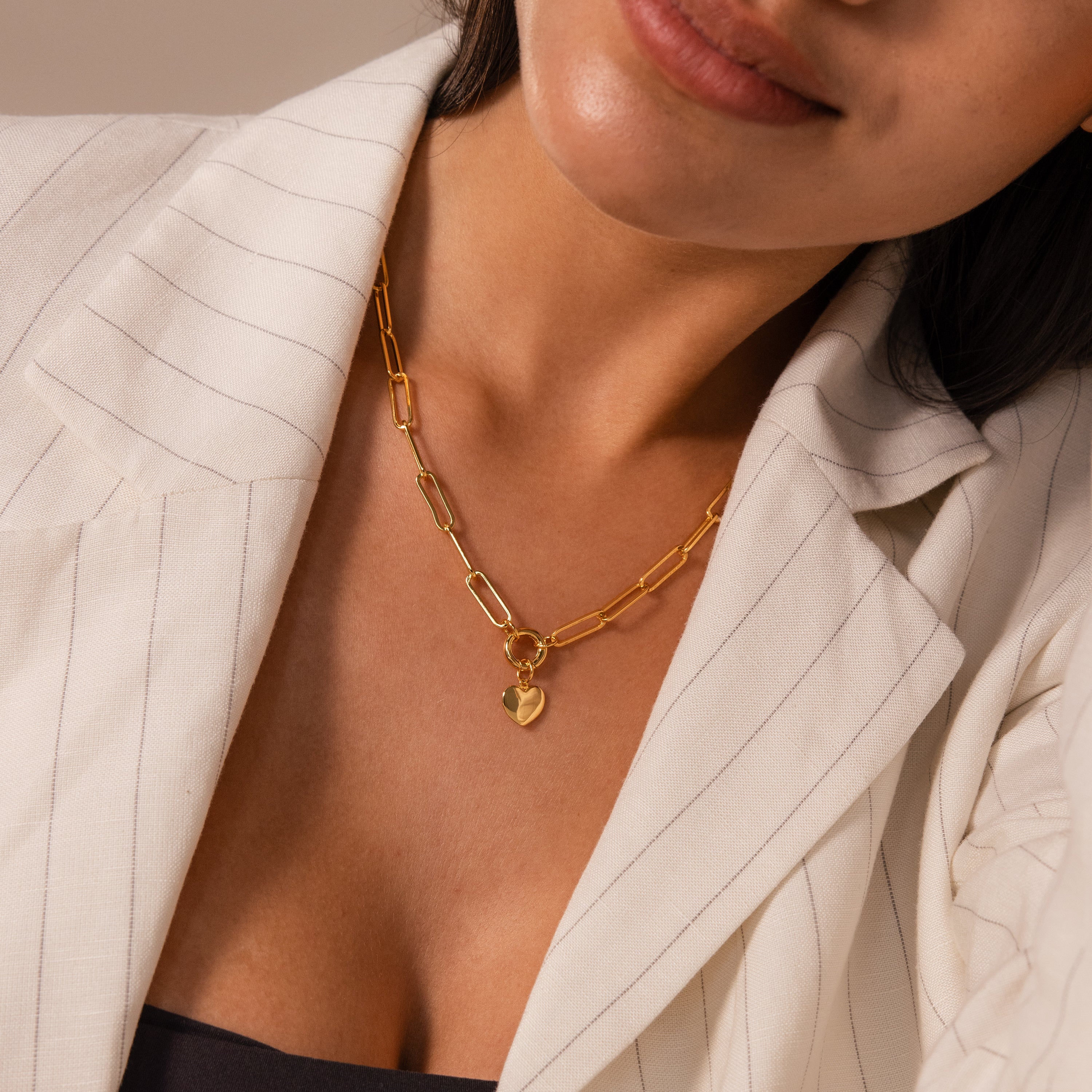 A woman in a white pinstripe blazer wears the Paperclip Heart Charm Necklace in 18K Gold, featuring a heart pendant.