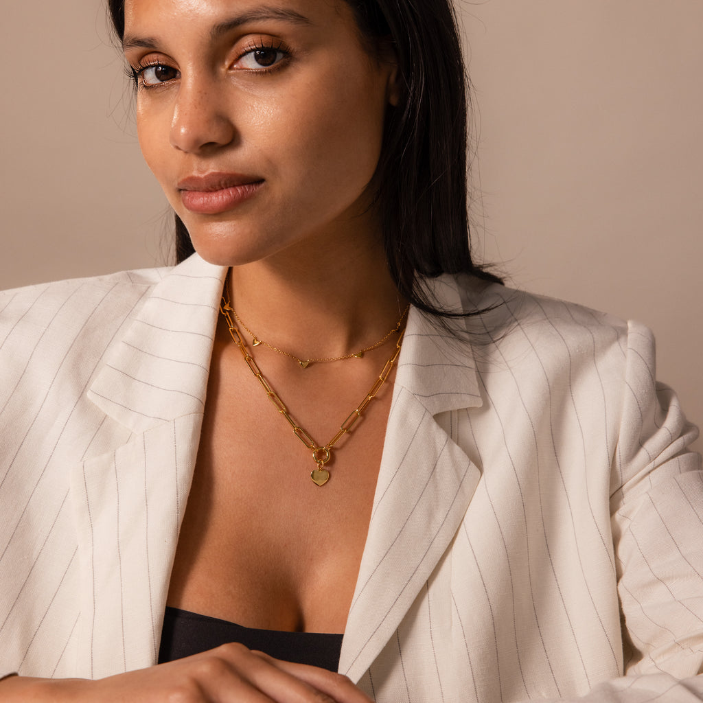 A woman in a white pinstripe blazer wears the Paperclip Heart Charm Necklace in 18K Gold, styled with layered gold chains, looking at the camera with a neutral expression.